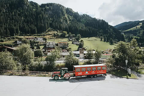 Der rote Großarltal-Express mit seinem Anhänger steht vor einer malerischen Landschaft aus Wäldern, Wiesen und Häusern. Gäste des Hotel Tauernhof in Großarl können mit diesem Zug die Umgebung bequem entdecken.