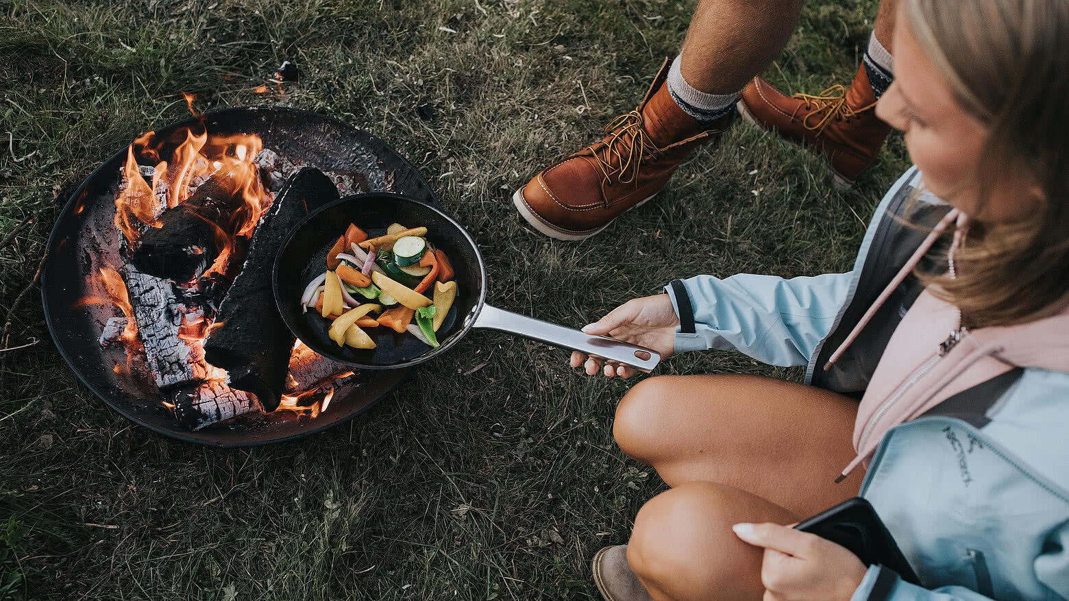 Eine junge Frau im Wanderoutfit legt eine gusseiserne Pfanne mit Gemüse auf einen Grill.