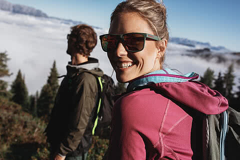 Zwei Wanderer in den Bergen nahe dem Hotel Tauernhof Großarl, eine Frau im Vordergrund mit Sonnenbrille und pinker Jacke lächelt in die Kamera, während die Landschaft von dichten Wäldern und Wolken umgeben ist.