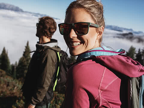 Eine junge Wanderin mit Sonnenbrille und einer pinken Jacke und einem Wanderrucksack lächelt in die Kamera. Im Hintergrund ist ihr Partner und eine Wolkendecke über den Bergen zu erkennen.