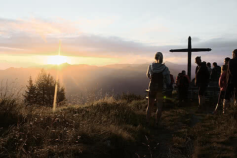 Wanderer genießen den Sonnenaufgang an einem Gipfelkreuz in den Alpen nahe dem Hotel Tauernhof Großarl, umgeben von beeindruckender Berglandschaft.