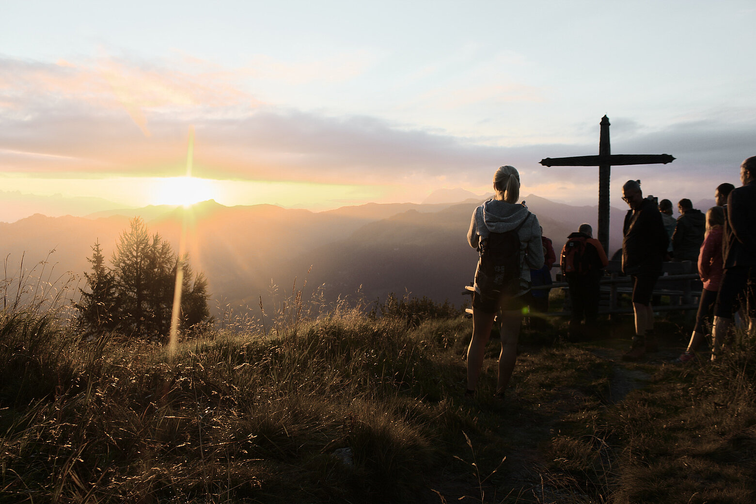 Wanderer genießen den Sonnenaufgang an einem Gipfelkreuz in den Alpen nahe dem Hotel Tauernhof Großarl, umgeben von beeindruckender Berglandschaft.