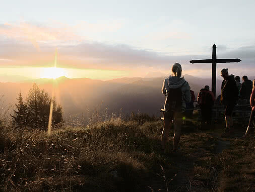Wanderer genießen den Sonnenaufgang an einem Gipfelkreuz in den Alpen nahe dem Hotel Tauernhof Großarl, umgeben von beeindruckender Berglandschaft.