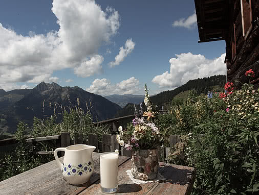 Holztisch vor der Maurachalm mit einer Kanne und einem Glas frischer Milch sowie einem Strauß bunter Wiesenblumen in einer Steinvase. Im Hintergrund sieht man ein Bergparonama.