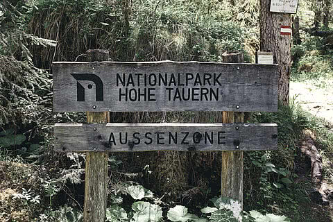 Ein hölzernes Schild markiert die Außenzone des Nationalparks Hohe Tauern, umgeben von dichter Waldlandschaft. Das Hotel Tauernhof in Großarl ist ein idealer Ausgangspunkt für Ausflüge in diese beeindruckende Naturregion.