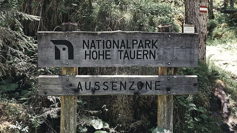 Ein hölzernes Schild markiert die Außenzone des Nationalparks Hohe Tauern, umgeben von dichter Waldlandschaft. Das Hotel Tauernhof in Großarl ist ein idealer Ausgangspunkt für Ausflüge in diese beeindruckende Naturregion.