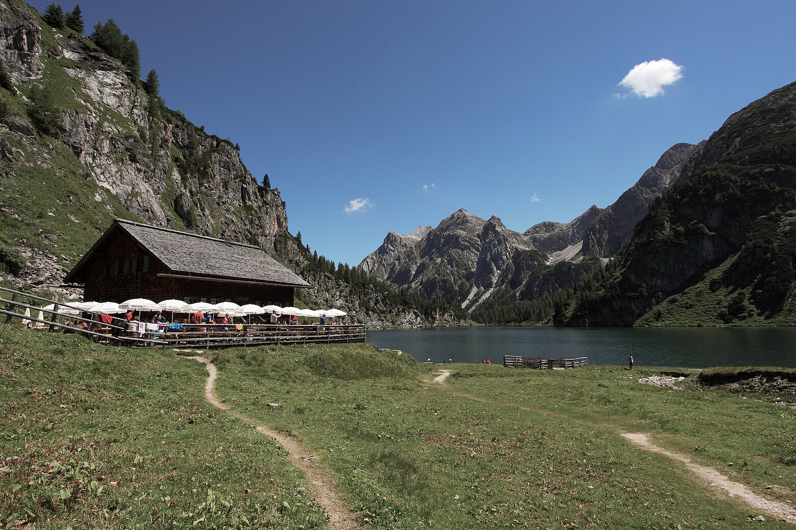 Die Tappenkarseehütte neben dem größten Gebirgssee der Ostalpen, dem Tappenkarsee. Im Hintergrund ist ein strahlend blauer Himmel und im Vordergrund satte Sommerwiesen. Auf der Terasse sitzen viele Gäste unter weißen Sonnenschirmen.