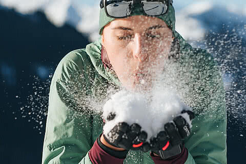 Frau in Winterkleidung pustet Schnee aus ihren Händen, umgeben von einer verschneiten Berglandschaft
