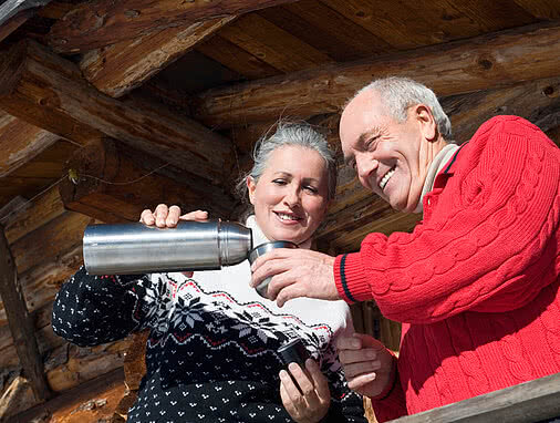 Älteres Paar steht auf sonnenbeschienenem Holzbalkon und gießt zufrieden lächelnd Tee aus Thermoskanne ein