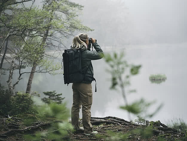 Frau mit Outdoor-Rucksack beobachtet die Natur mit Fernglas an einem nebligen See im Alpen Retreat Urlaub