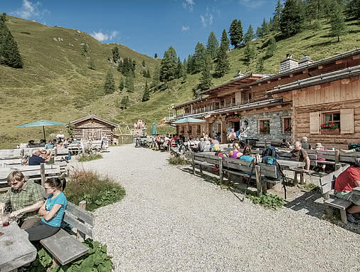 Sonnenterrasse der Loosbühelalm im Großarltal mit vielen Gästen an rustikalen Holzbänken. Im Hintergrund grüne Almhänge mit einzelnen Bäumen.