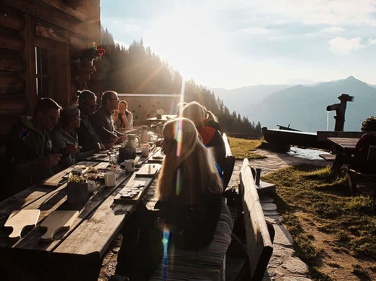Gäste genießen ein Essen auf der Sonnenterrasse einer Berghütte nahe Hotel Tauernhof Großarl, mit Blick auf die Alpenlandschaft bei Sonnenuntergang.