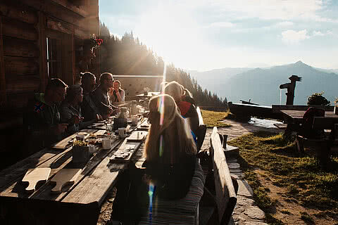 Gäste genießen ein Essen auf der Sonnenterrasse einer Berghütte nahe Hotel Tauernhof Großarl, mit Blick auf die Alpenlandschaft bei Sonnenuntergang.