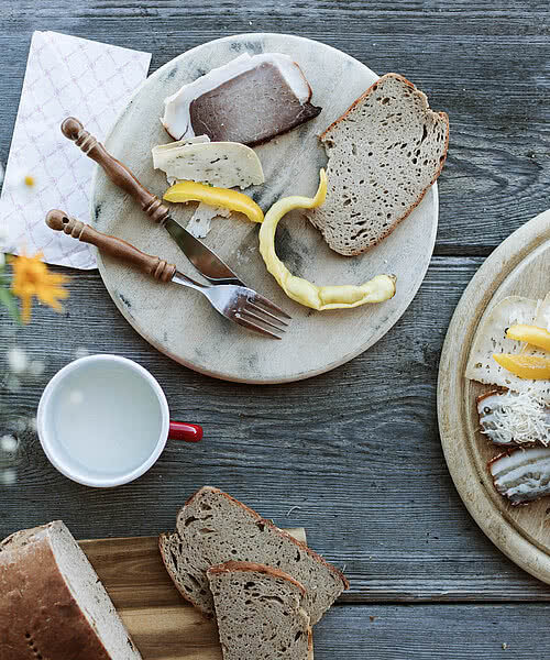 Zwei Holzplatten mit einer köstlichen Jause aus Käse, Wurst und frischem Brot, umgeben von einer Tasse und Blumenarrangement.