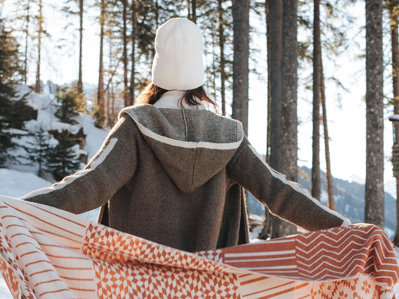 Frau in Winterkleidung mit ausgestreckten Armen im verschneiten Wald, umgeben von Sonnenschein und Natur