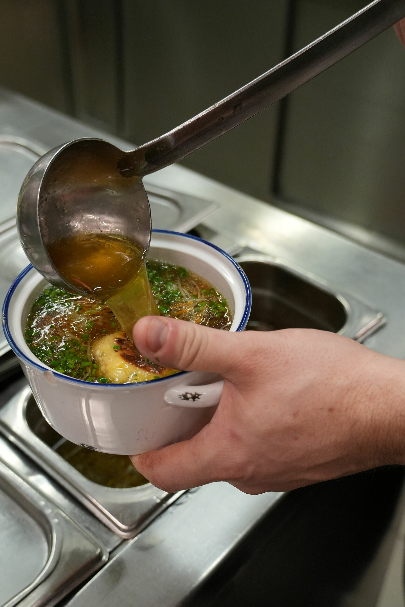 Ein Kopf schöpft warme Suppe in einen kleinen Emailletopf mit zwei Kaspressknödel