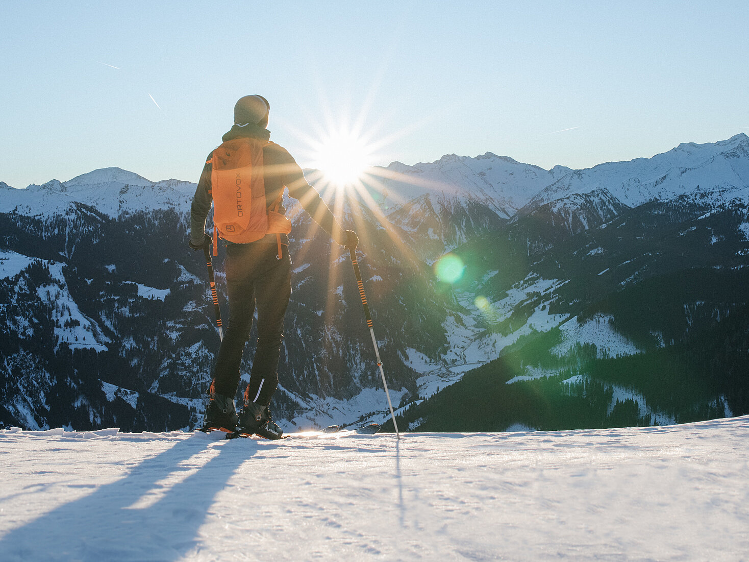 Skitourengeher mit orangefarbenem Rucksack genießt den Sonnenaufgang über verschneiten Bergen