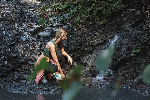 Wanderin in sportlicher Kleidung kniet an einem kleinen Gebirgsbach nahe dem Hotel Tauernhof Großarl, umgeben von einer felsigen und grünen Naturkulisse.