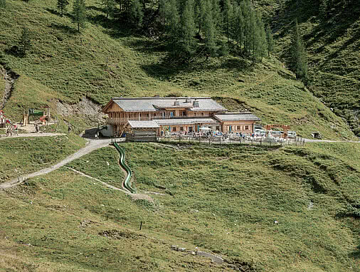 Die Loosbühelalm im Großarltal: eine große Hütte inmitten grüner Almwiesen, mit Sonnenterrasse. Links daneben befindet sich ein Kinderspielplatz mit Rutsche und Schaukeln.