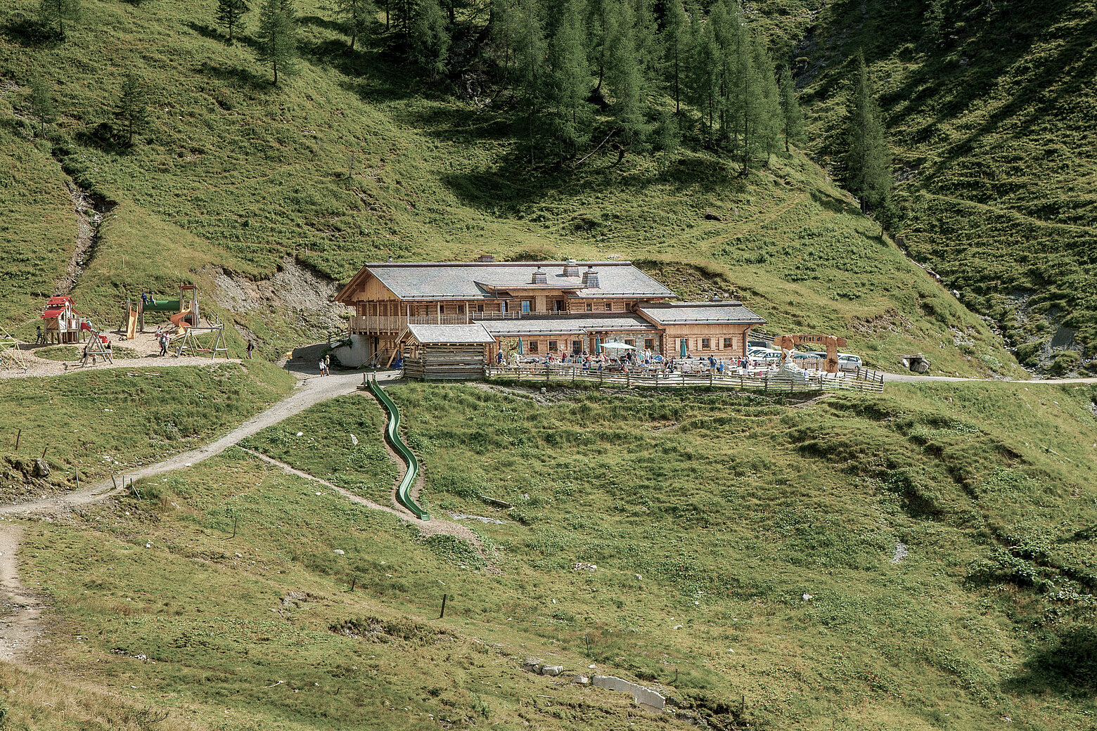 Die Loosbühelalm im Großarltal: eine große Hütte inmitten grüner Almwiesen, mit Sonnenterrasse. Links daneben befindet sich ein Kinderspielplatz mit Rutsche und Schaukeln.