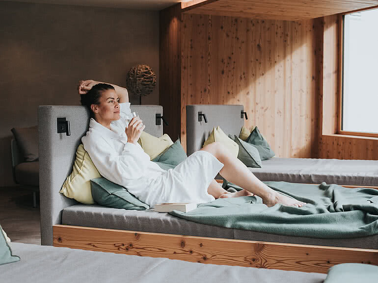 Entspannte Frau im Bademantel beim Alpen Retreat, gemütlich auf einer Liege in einem modernen Wellnessbereich, umgeben von Holzvertäfelung