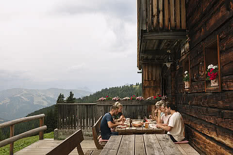 Eine Gruppe von fünf Personen genießt ein gemütliches Mittagessen auf einer rustikalen Terrasse in den Bergen.